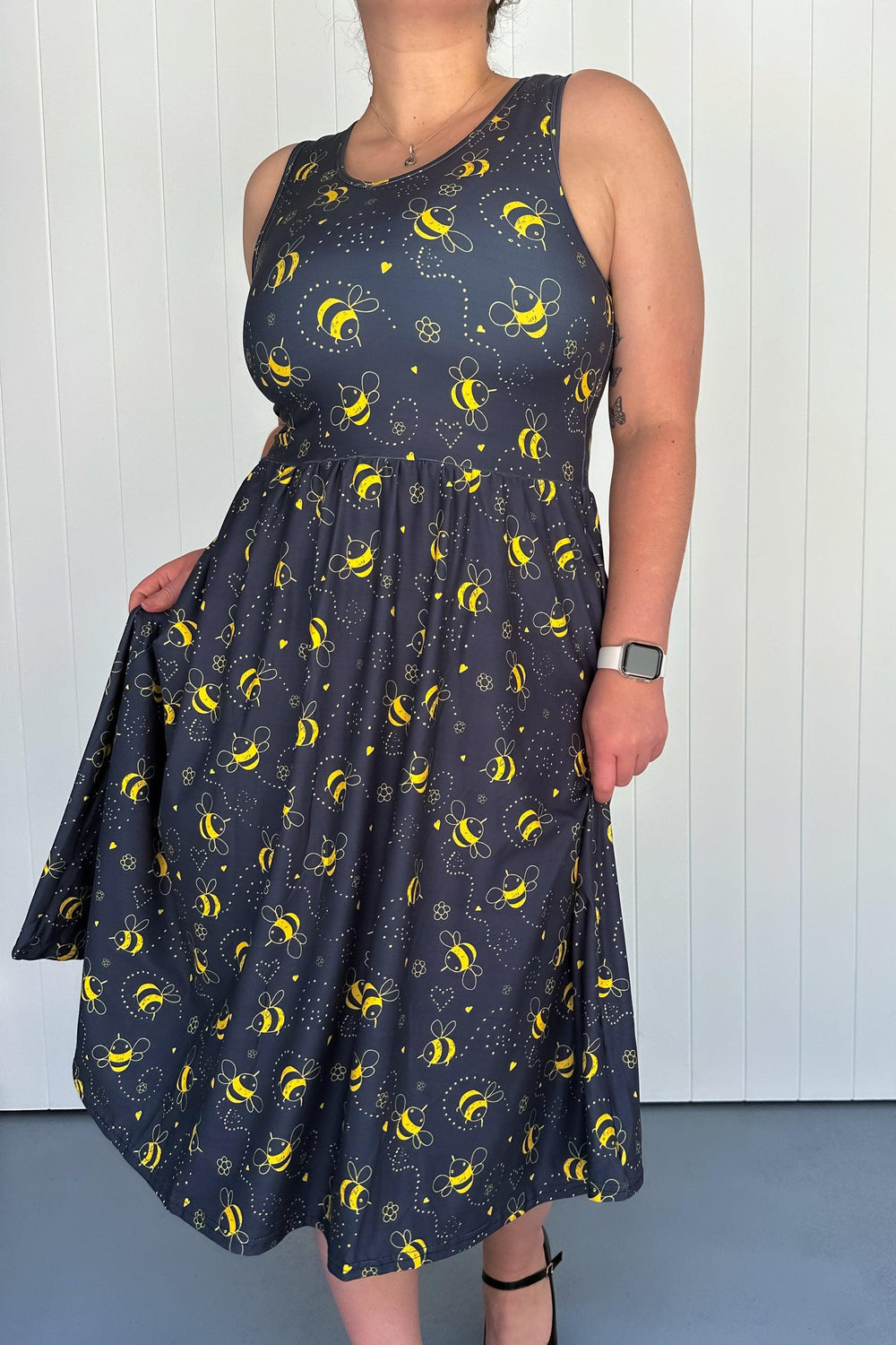 A sleeveless midi dress with a crew neckline, featuring a charcoal-grey background with a pattern of yellow bees. The dress has deep side pockets and appears to be made of a stretchy fabric.