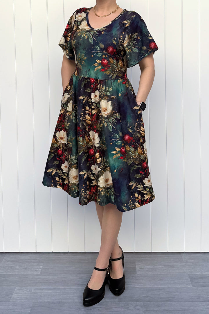 Person wearing a dark Christmas-themed floral dress with white flowers and red berries, standing against a white wooden panel background