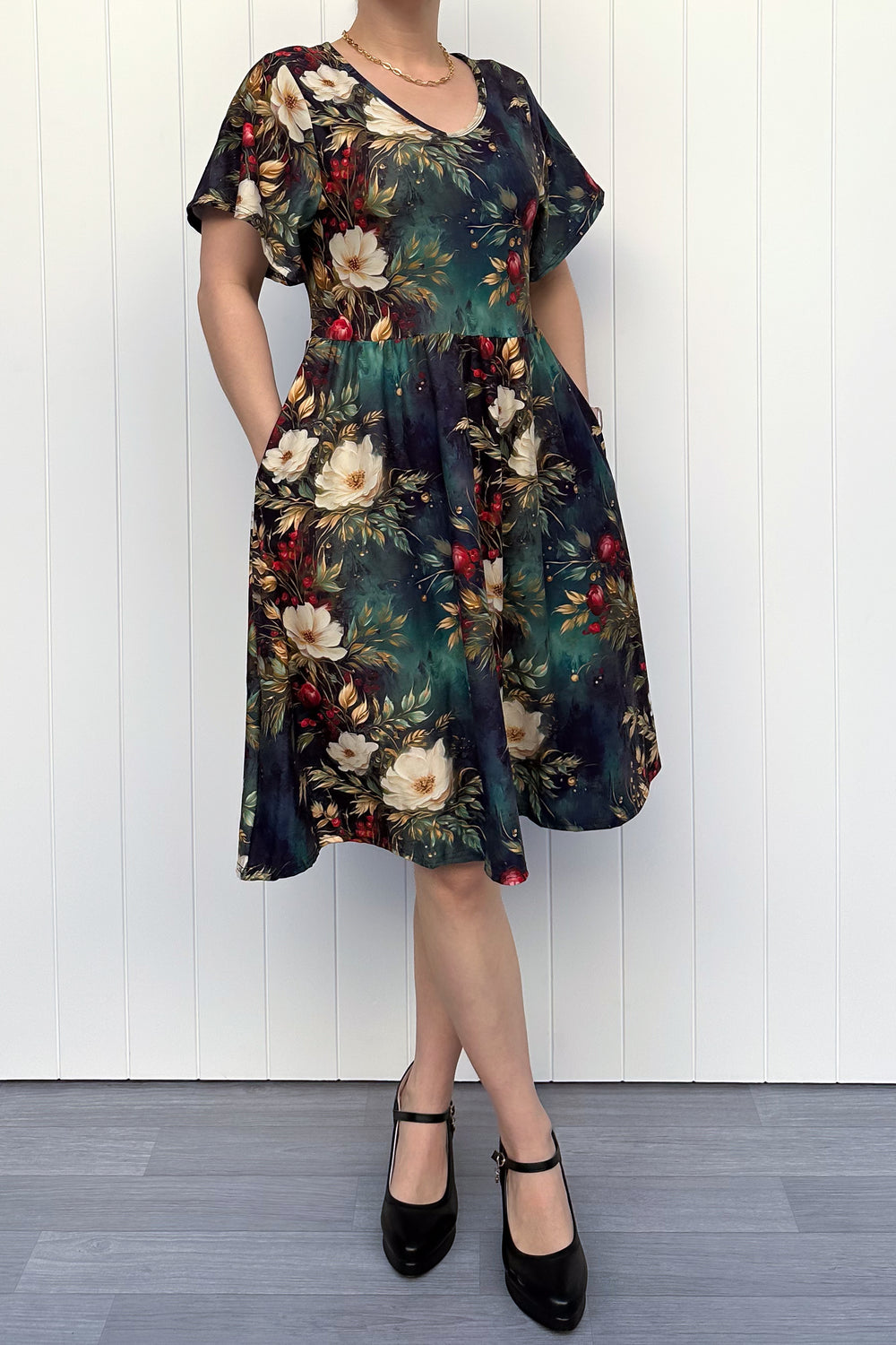Person wearing a dark Christmas-themed floral dress with white flowers and red berries, standing against a white wooden panel background