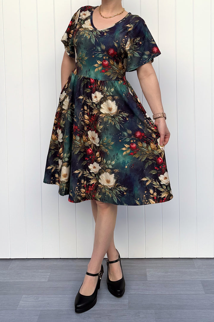 Person wearing a dark Christmas-themed floral dress with white flowers and red berries, standing against a white wooden panel background