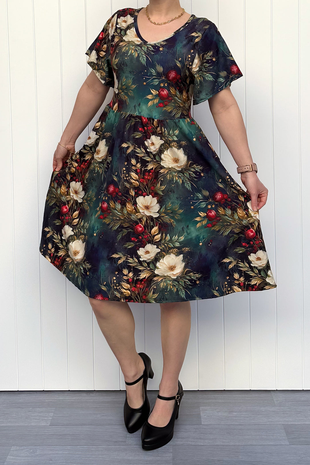 Person wearing a dark Christmas-themed floral dress with white flowers and red berries, standing against a white wooden panel background