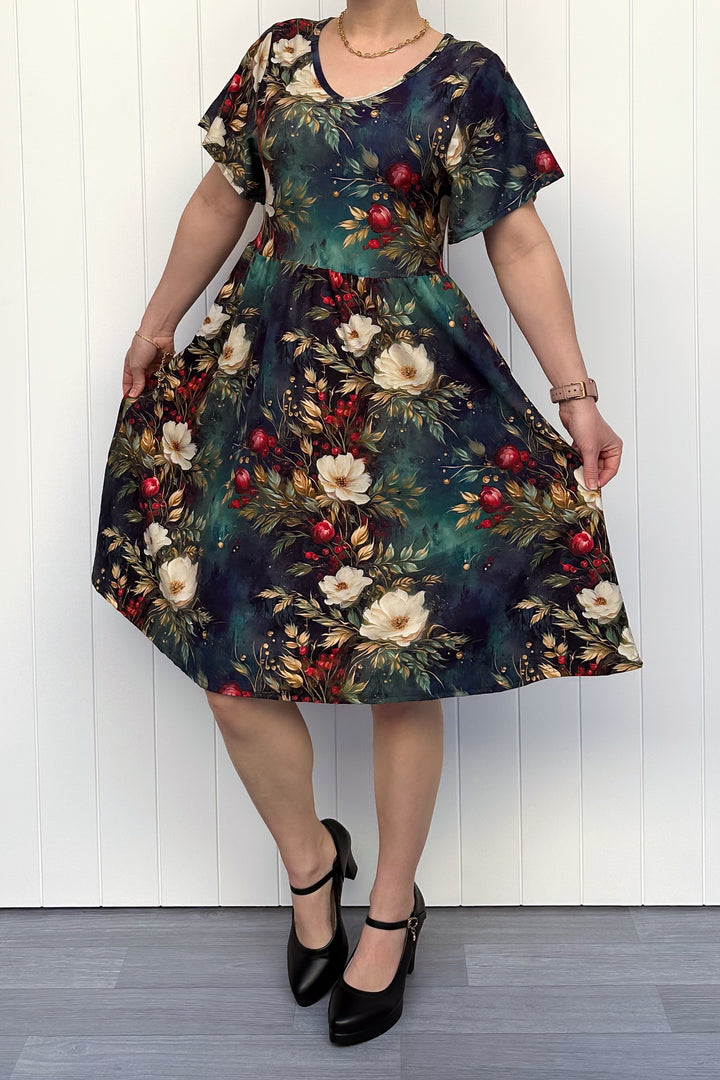 Person wearing a dark Christmas-themed floral dress with white flowers and red berries, standing against a white wooden panel background