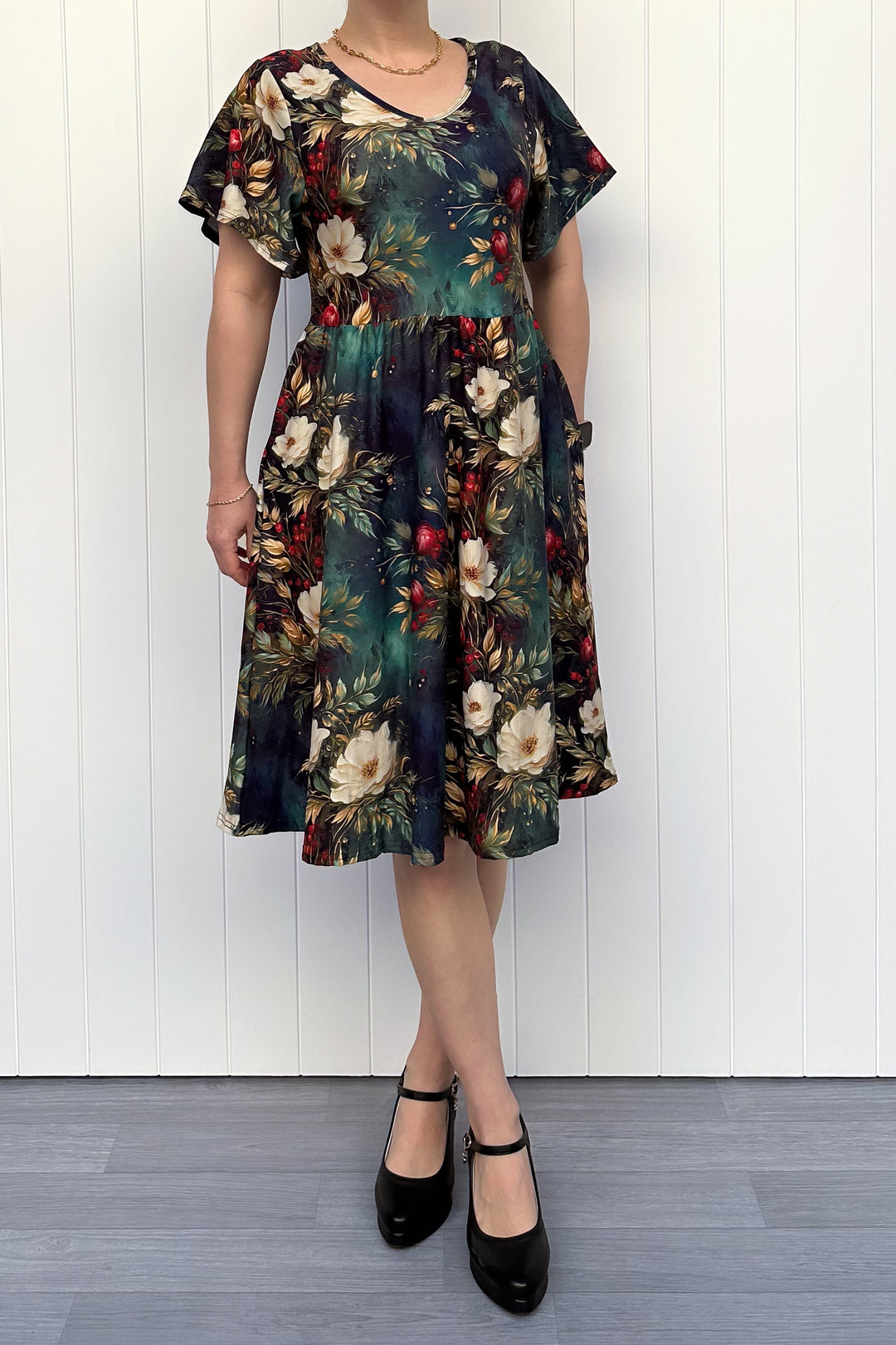 Person wearing a dark Christmas-themed floral dress with white flowers and red berries, standing against a white wooden panel background