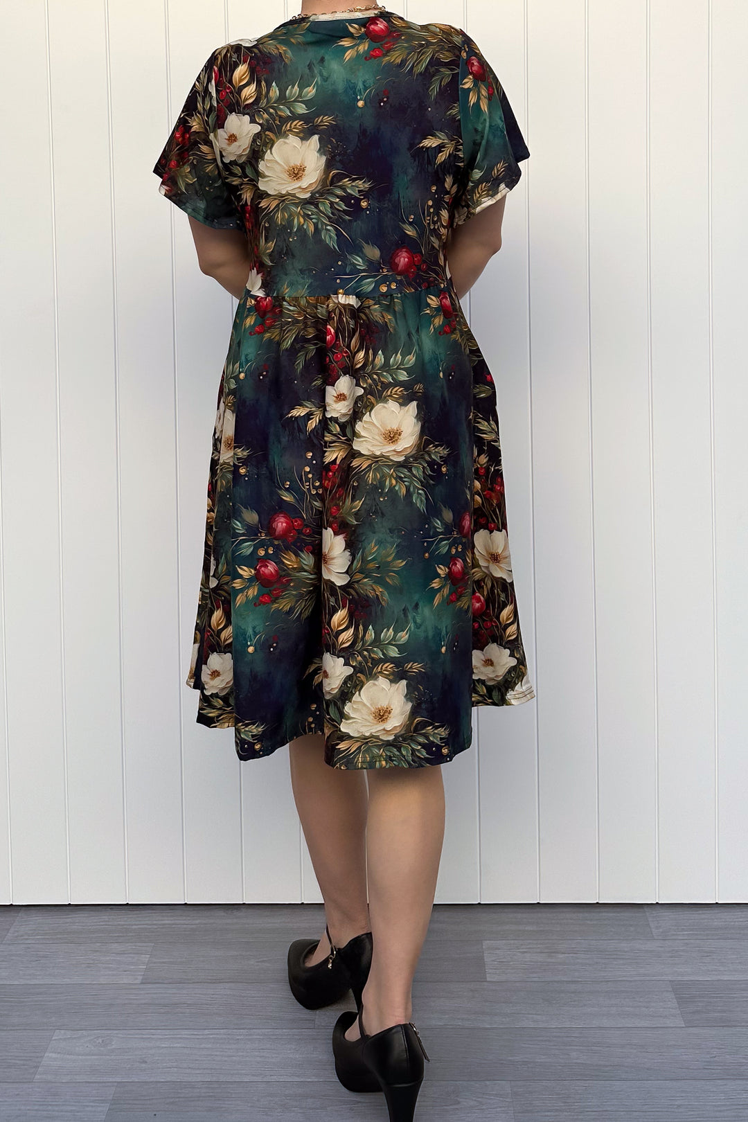 Person wearing a dark Christmas-themed floral dress with white flowers and red berries, standing against a white wooden panel background