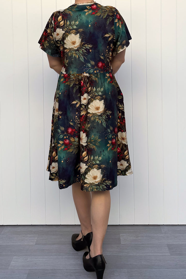 Person wearing a dark Christmas-themed floral dress with white flowers and red berries, standing against a white wooden panel background