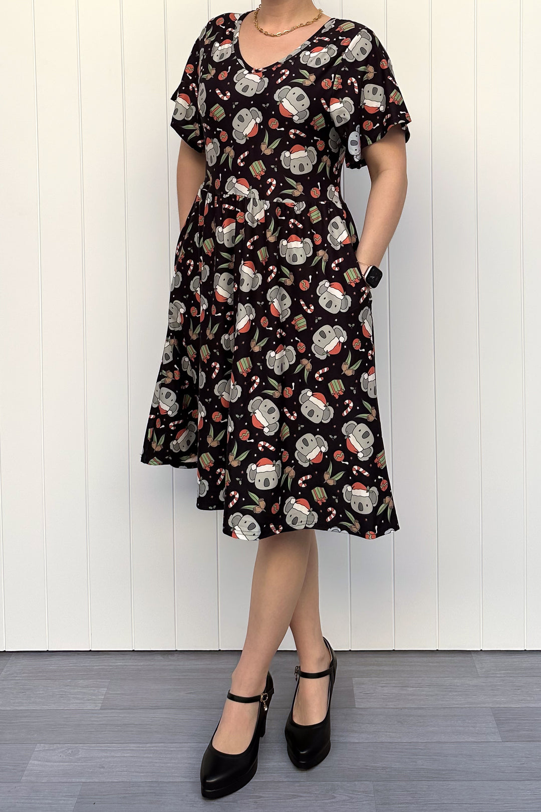 Person wearing a dress featuring a pattern of koalas wearing santa hats and candy canes on a black background, standing against a white wall.