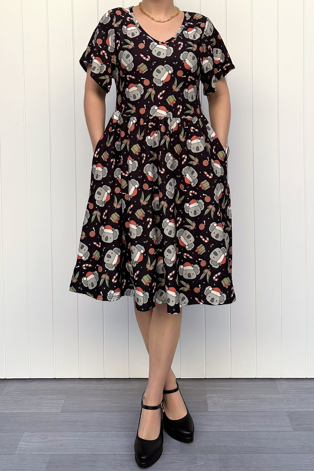 Person wearing a dress featuring a pattern of koalas wearing santa hats and candy canes on a black background, standing against a white wall.