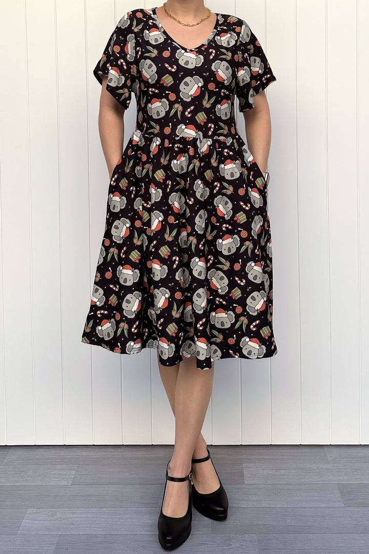Person wearing a dress featuring a pattern of koalas wearing santa hats and candy canes on a black background, standing against a white wall.