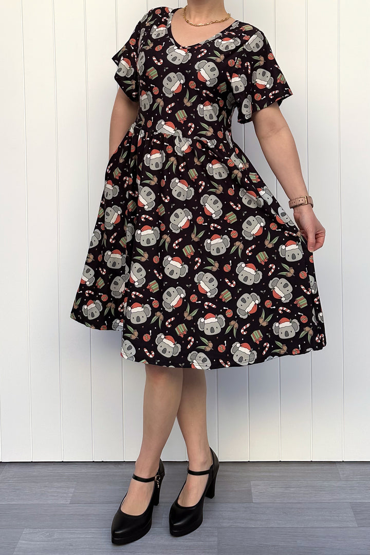 Person wearing a dress featuring a pattern of koalas wearing santa hats and candy canes on a black background, standing against a white wall.