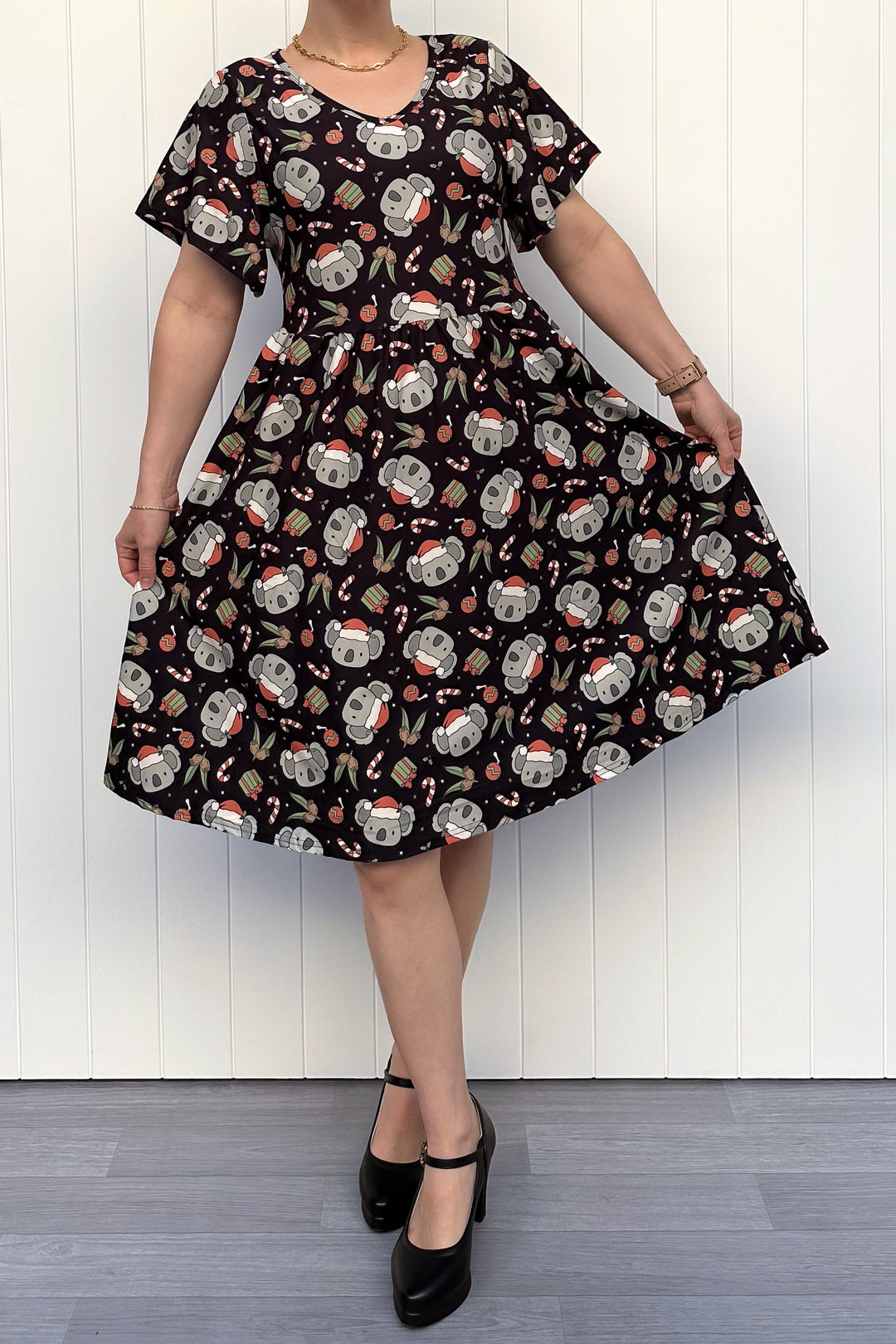 Person wearing a dress featuring a pattern of koalas wearing santa hats and candy canes on a black background, standing against a white wall.