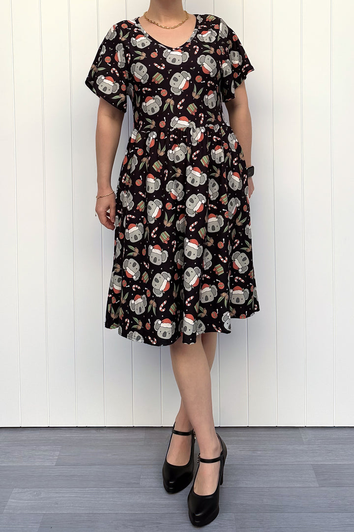 Person wearing a dress featuring a pattern of koalas wearing santa hats and candy canes on a black background, standing against a white wall.