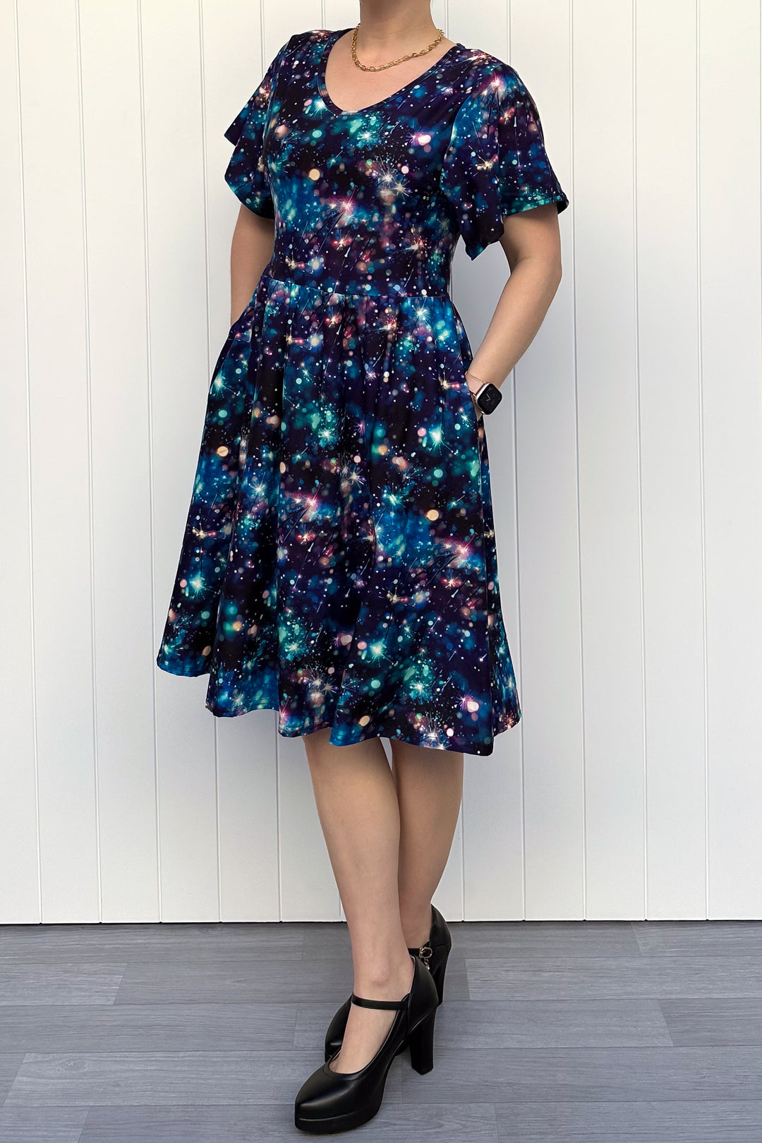 Person wearing a celebration sparkler-inspired patterned dress in blues, greens and pinks, standing against a white wall.