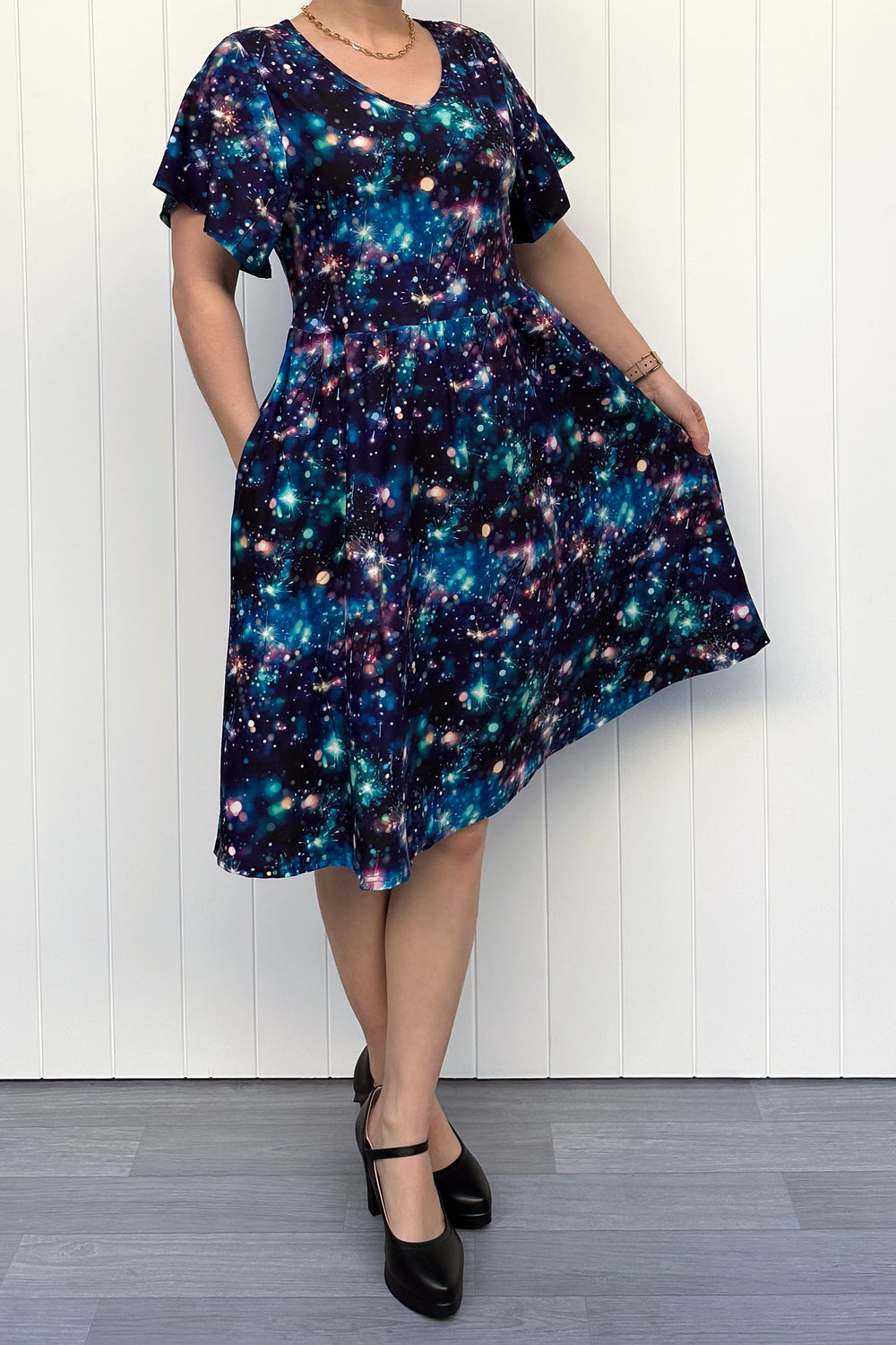 Person wearing a celebration sparkler-inspired patterned dress in blues, greens and pinks, standing against a white wall.
