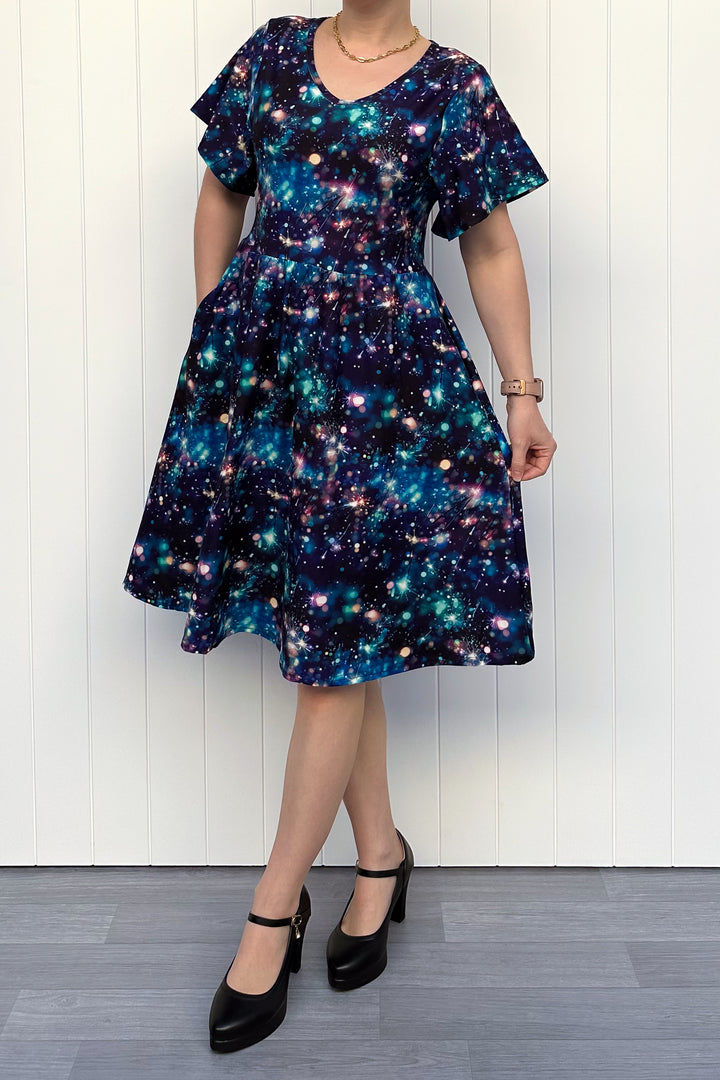Person wearing a celebration sparkler-inspired patterned dress in blues, greens and pinks, standing against a white wall.