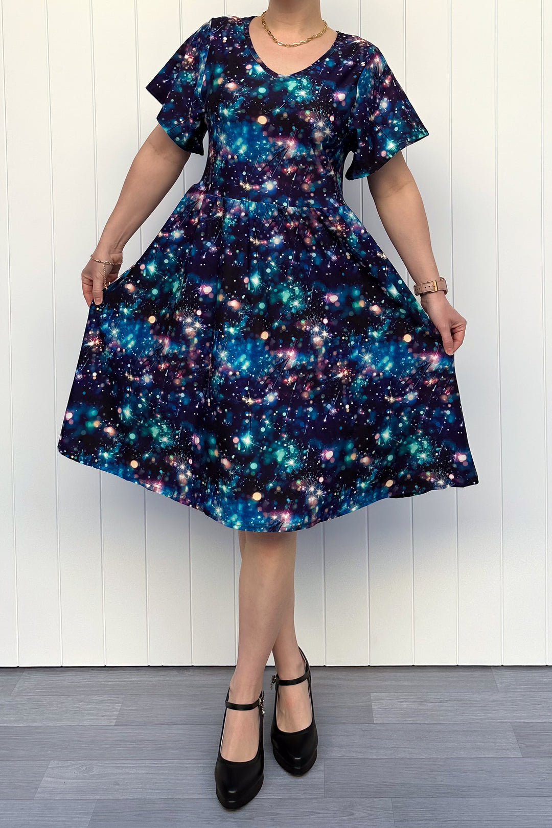 Person wearing a celebration sparkler-inspired patterned dress in blues, greens and pinks, standing against a white wall.