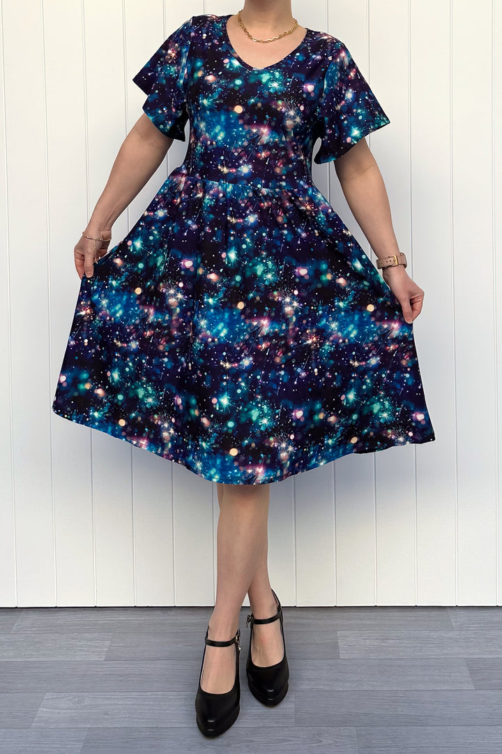 Person wearing a celebration sparkler-inspired patterned dress in blues, greens and pinks, standing against a white wall.