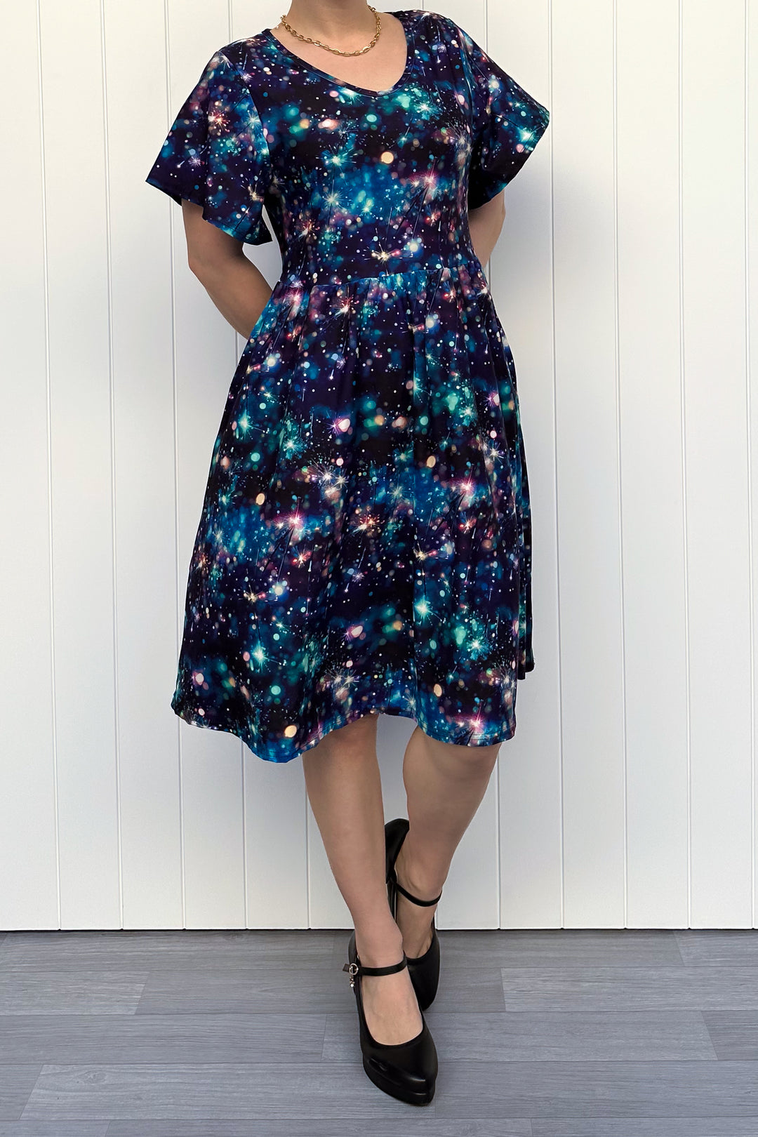 Person wearing a celebration sparkler-inspired patterned dress in blues, greens and pinks, standing against a white wall.