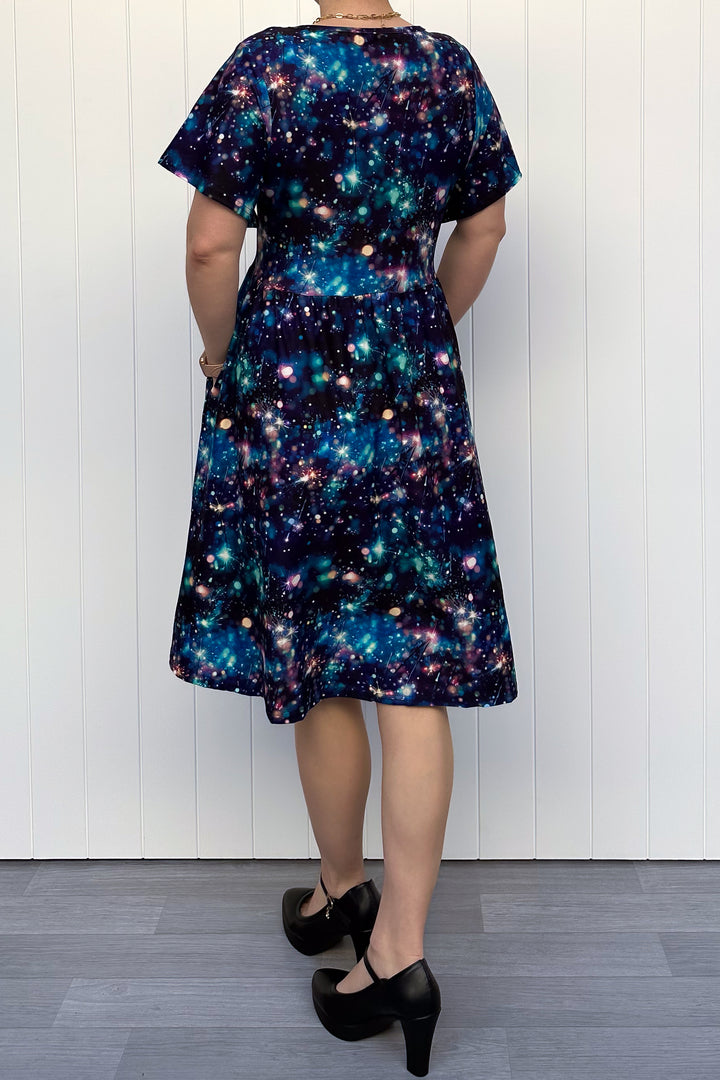 Person wearing a celebration sparkler-inspired patterned dress in blues, greens and pinks, standing against a white wall.