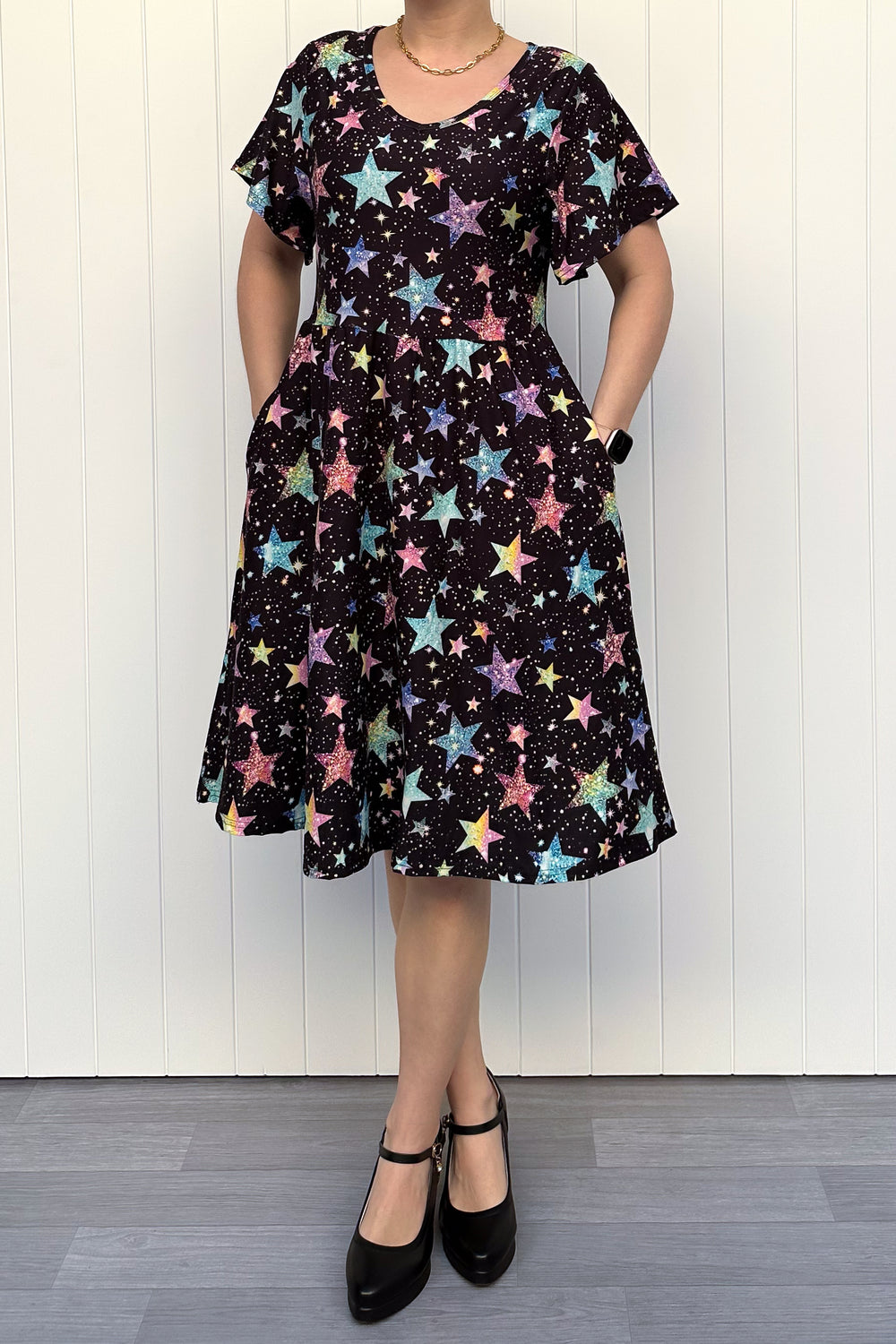 Person wearing a black dress with colourful sparkle-like star pattern, standing against a white wall.