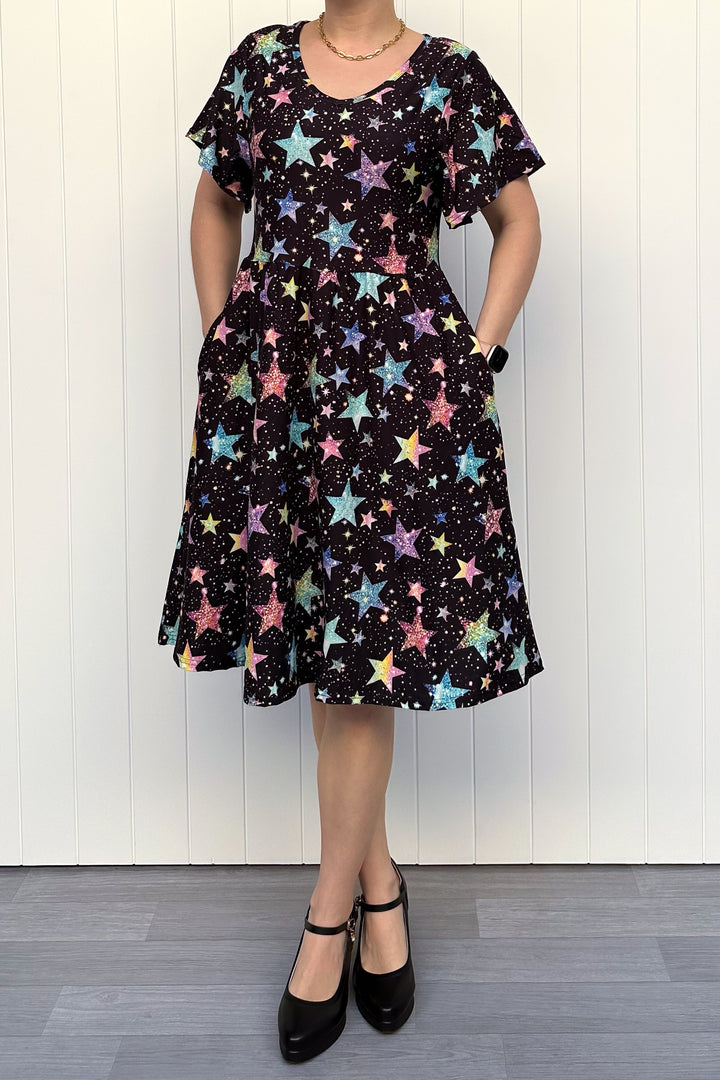 Person wearing a black dress with colourful sparkle-like star pattern, standing against a white wall.