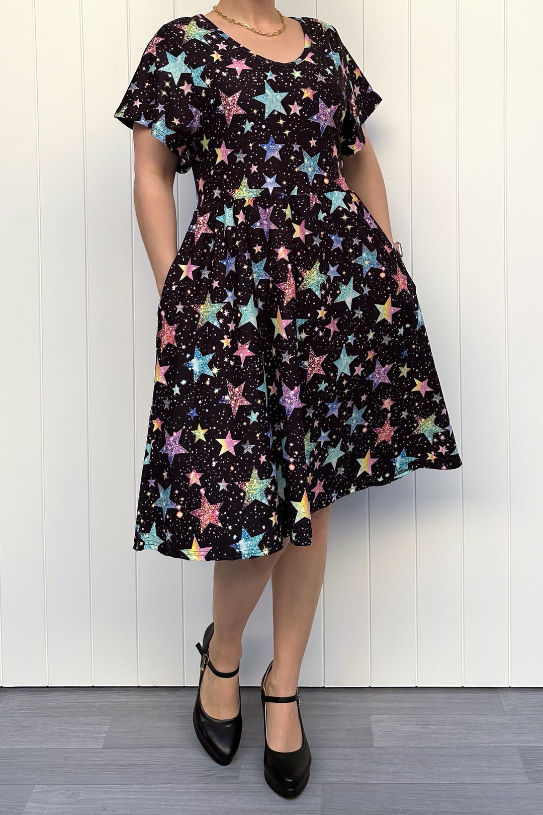 Person wearing a black dress with colourful sparkle-like star pattern, standing against a white wall.