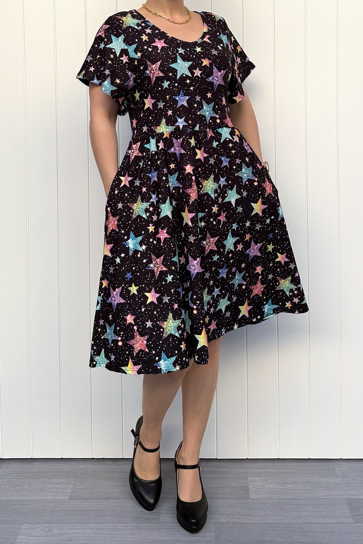 Person wearing a black dress with colourful sparkle-like star pattern, standing against a white wall.