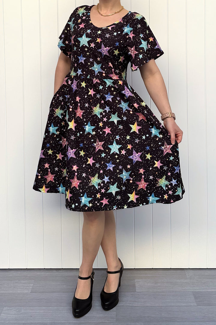 Person wearing a black dress with colourful sparkle-like star pattern, standing against a white wall.