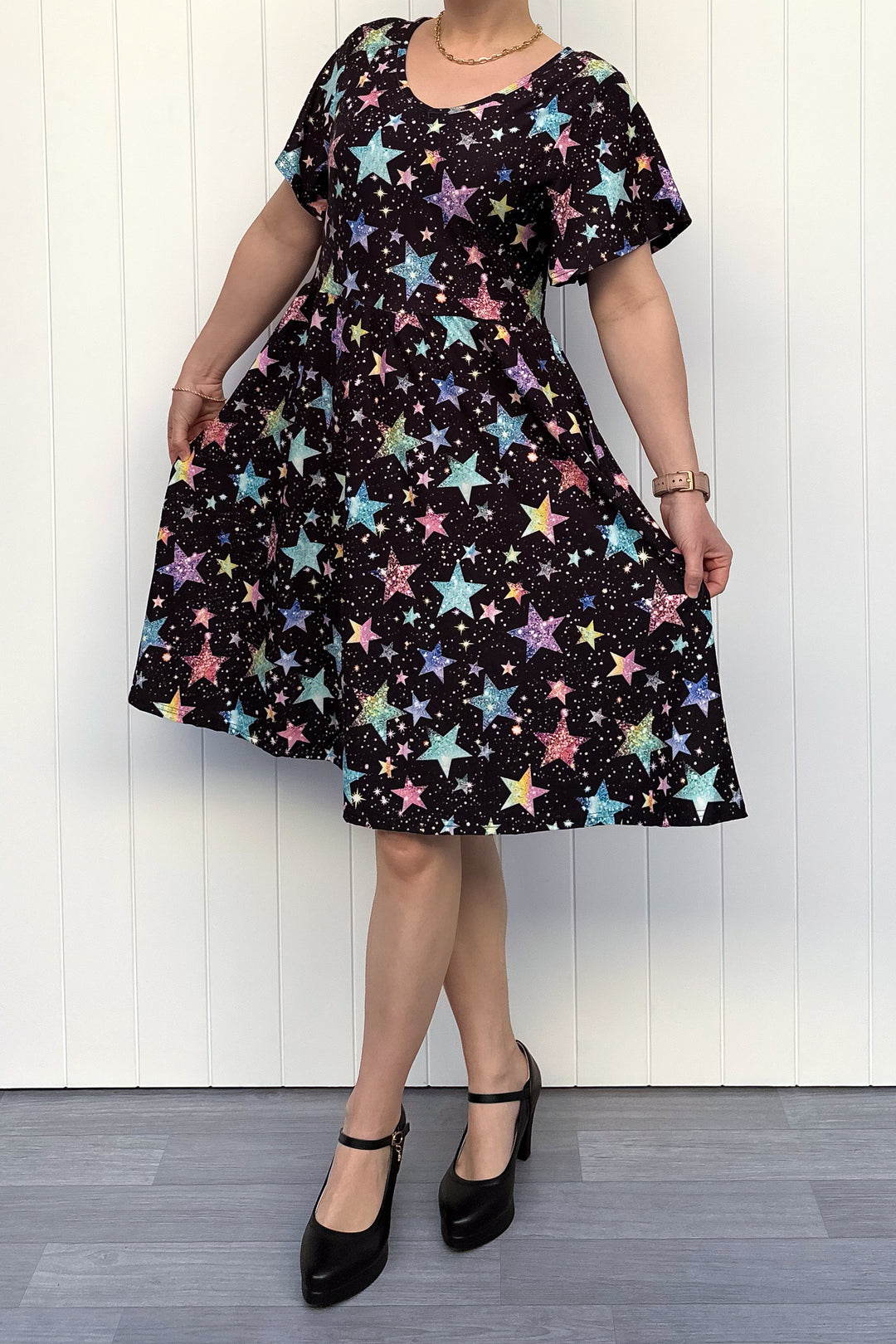 Person wearing a black dress with colourful sparkle-like star pattern, standing against a white wall.