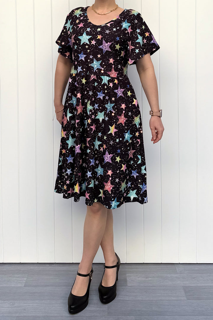 Person wearing a black dress with colourful sparkle-like star pattern, standing against a white wall.
