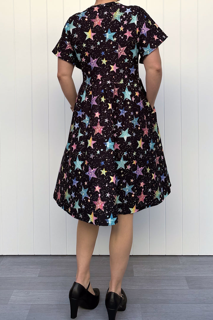 Person wearing a black dress with colourful sparkle-like star pattern, standing against a white wall.