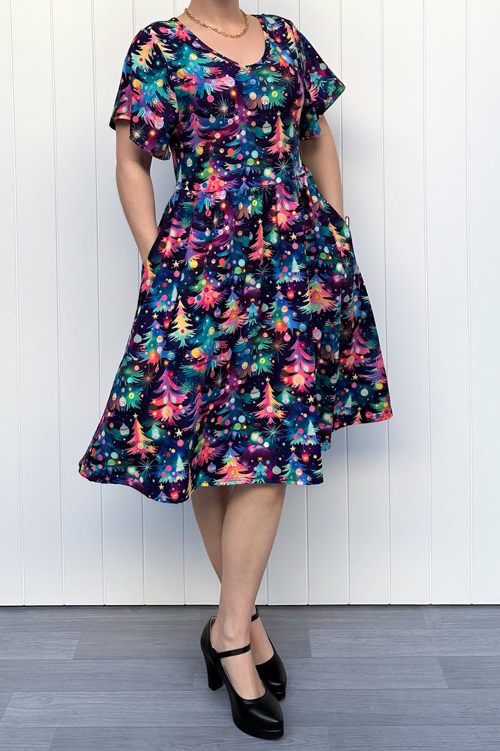 Person wearing a dress with a pattern of colourful Christmas trees against a white wooden panel background.