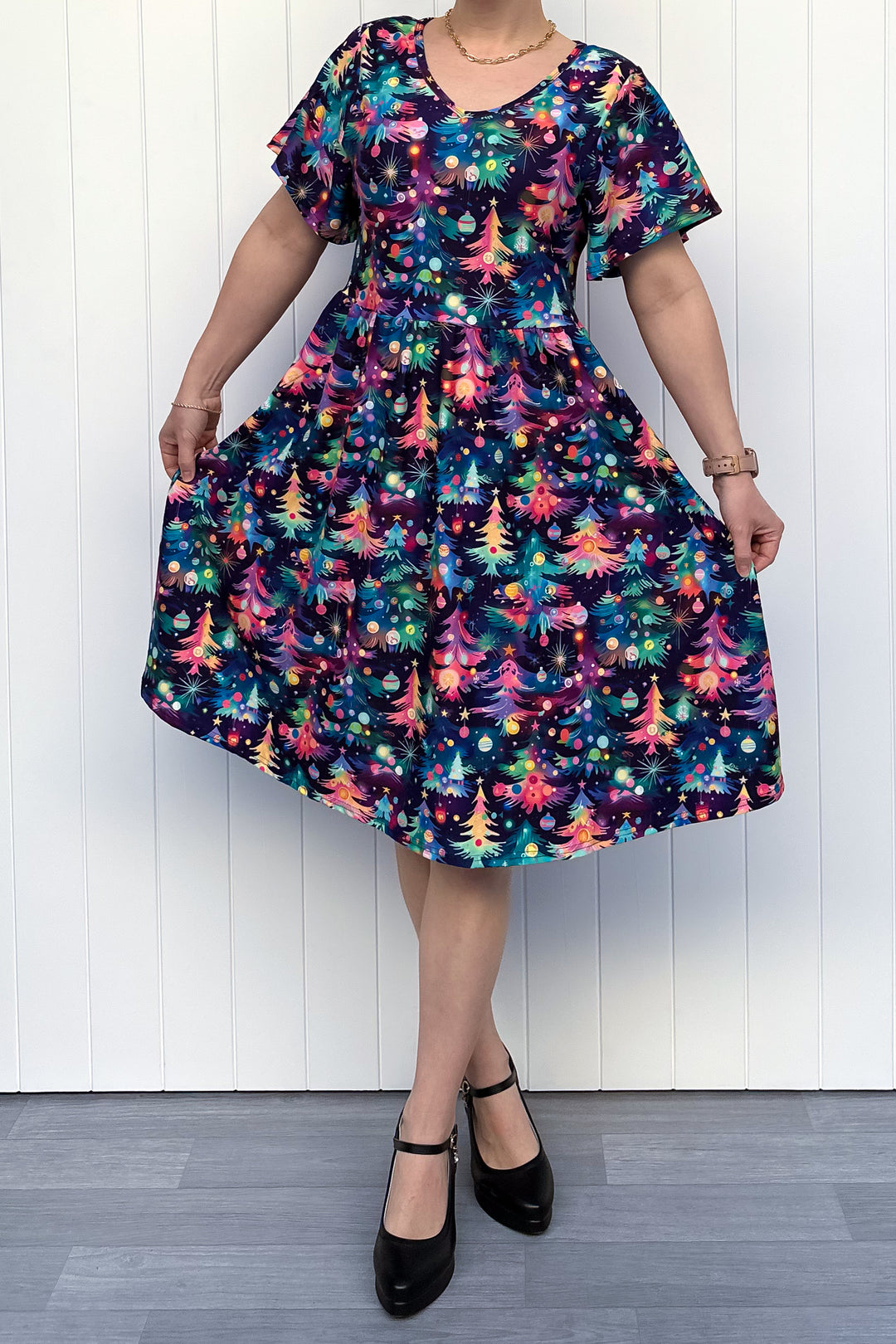 Person wearing a dress with a pattern of colourful Christmas trees against a white wooden panel background.