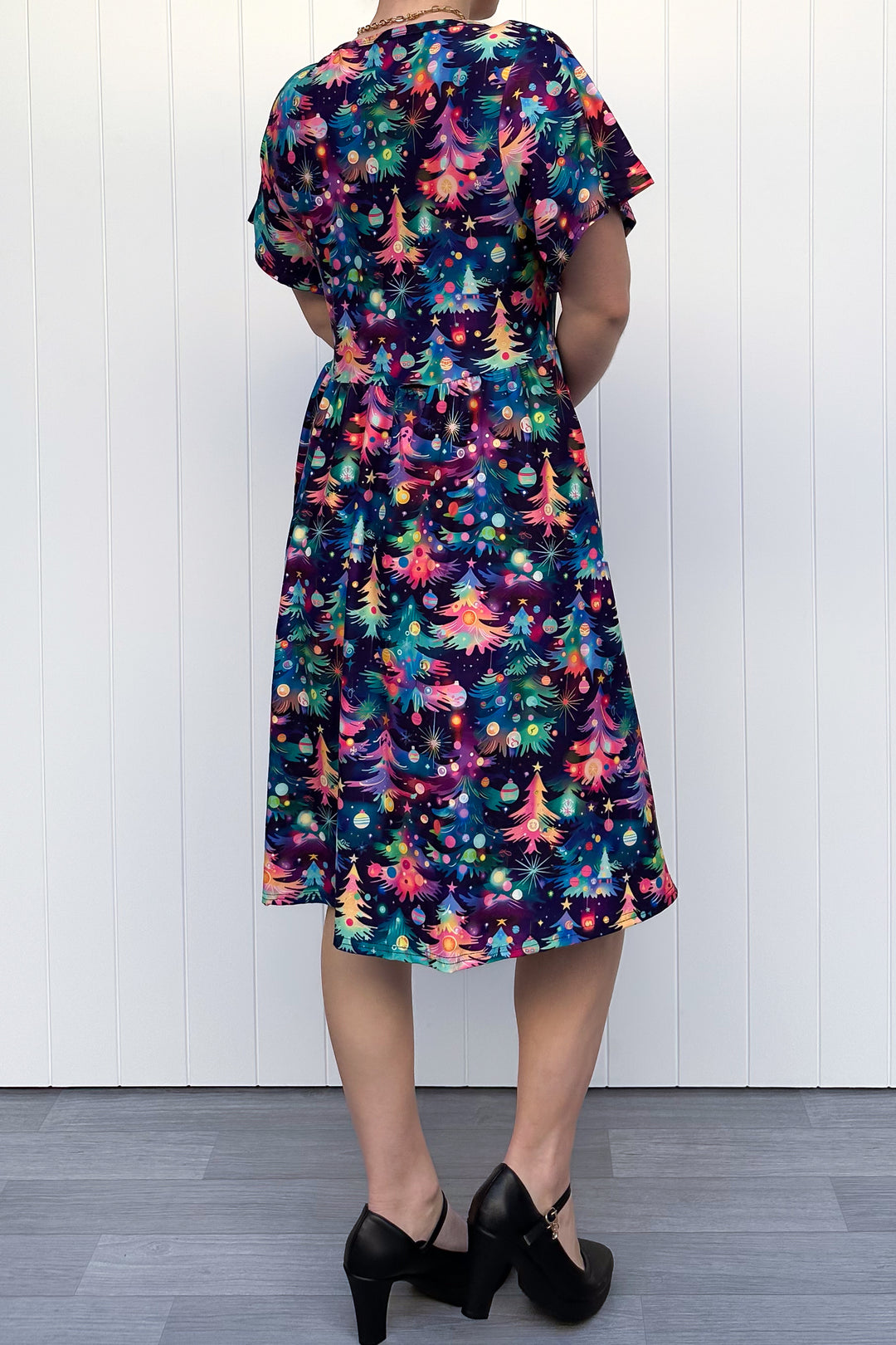 Person wearing a dress with a pattern of colourful Christmas trees against a white wooden panel background.