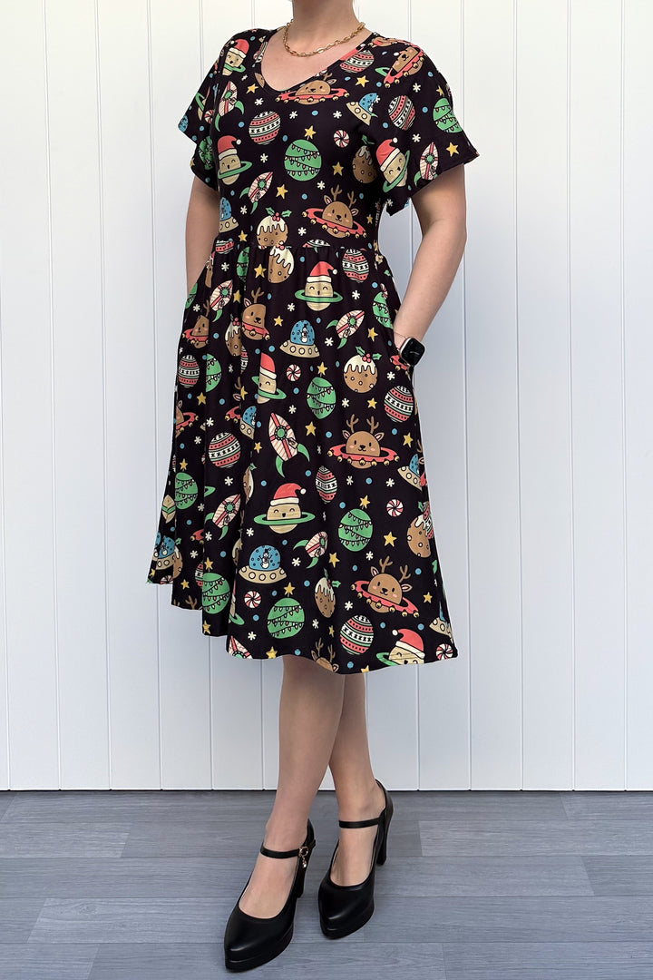 Person wearing a black dress with colorful space-themed Christmas pattern standing against a white wall.