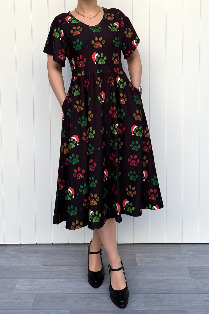 Person wearing a black dress with red, green and yellow paw print pattern with santa hats, standing against a white wooden panel background