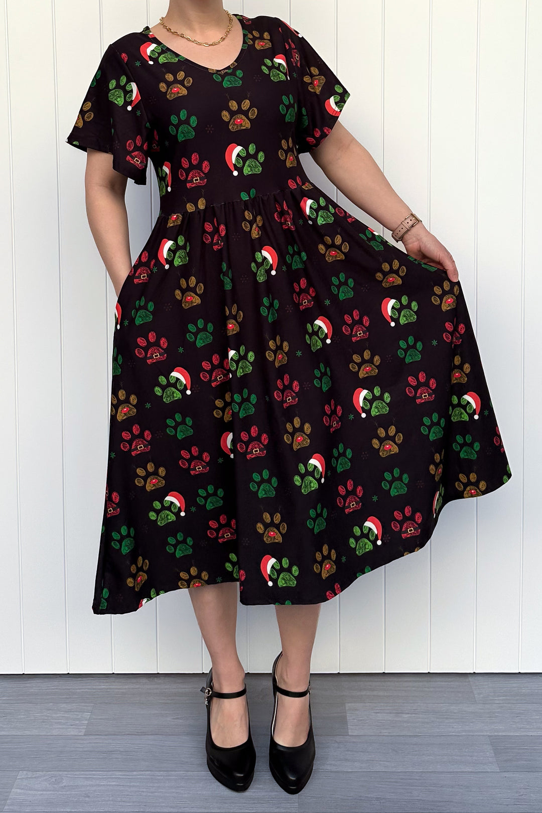 Person wearing a black dress with red, green and yellow paw print pattern with santa hats, standing against a white wooden panel background
