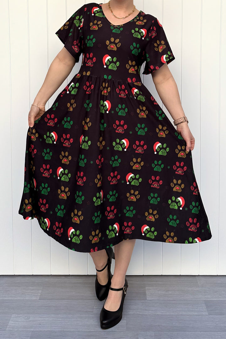 Person wearing a black dress with red, green and yellow paw print pattern with santa hats, standing against a white wooden panel background