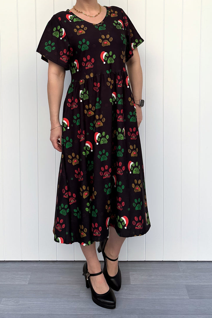 Person wearing a black dress with red, green and yellow paw print pattern with santa hats, standing against a white wooden panel background