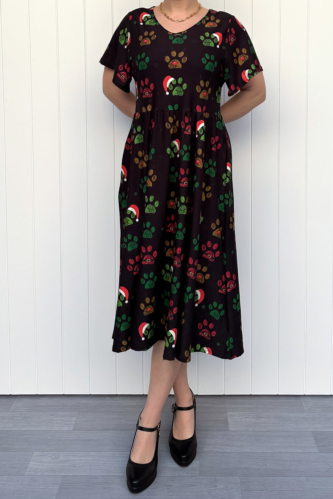 Person wearing a black dress with red, green and yellow paw print pattern with santa hats, standing against a white wooden panel background