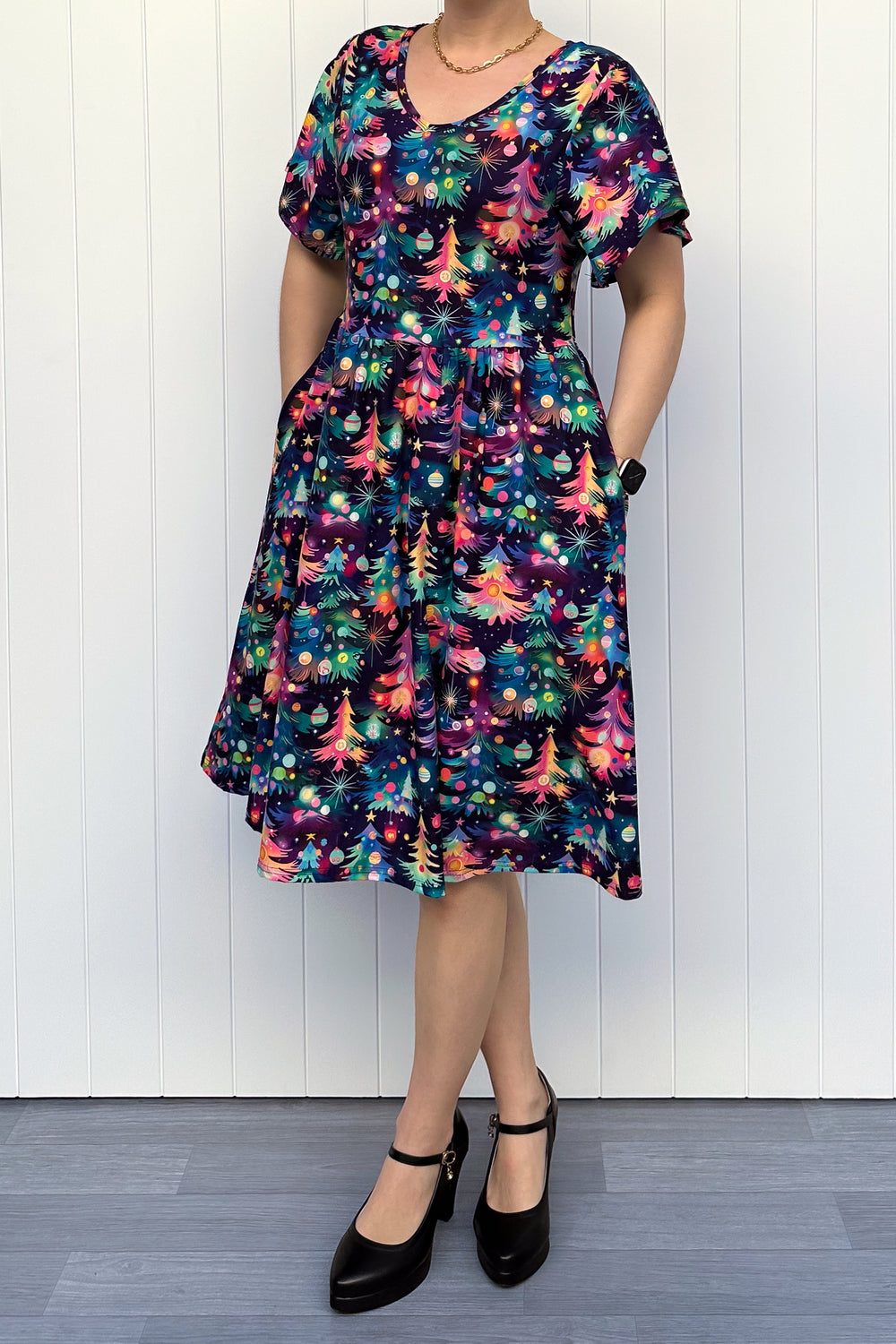 Person wearing a dress with a pattern of colourful Christmas trees against a white wooden panel background.