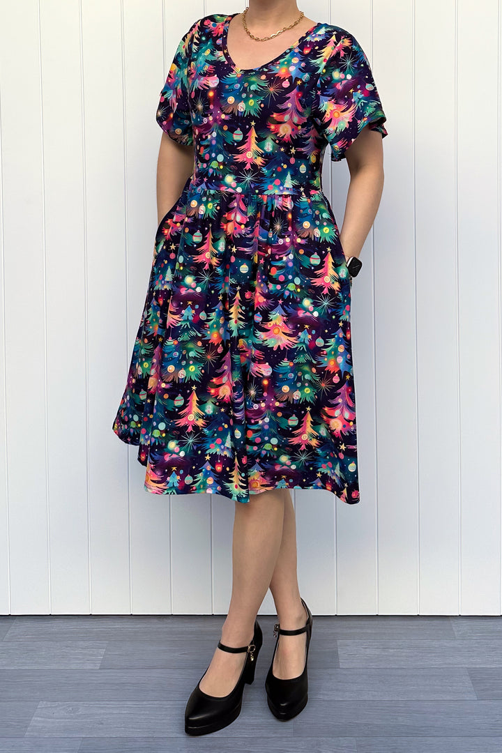 Person wearing a dress with a pattern of colourful Christmas trees against a white wooden panel background.