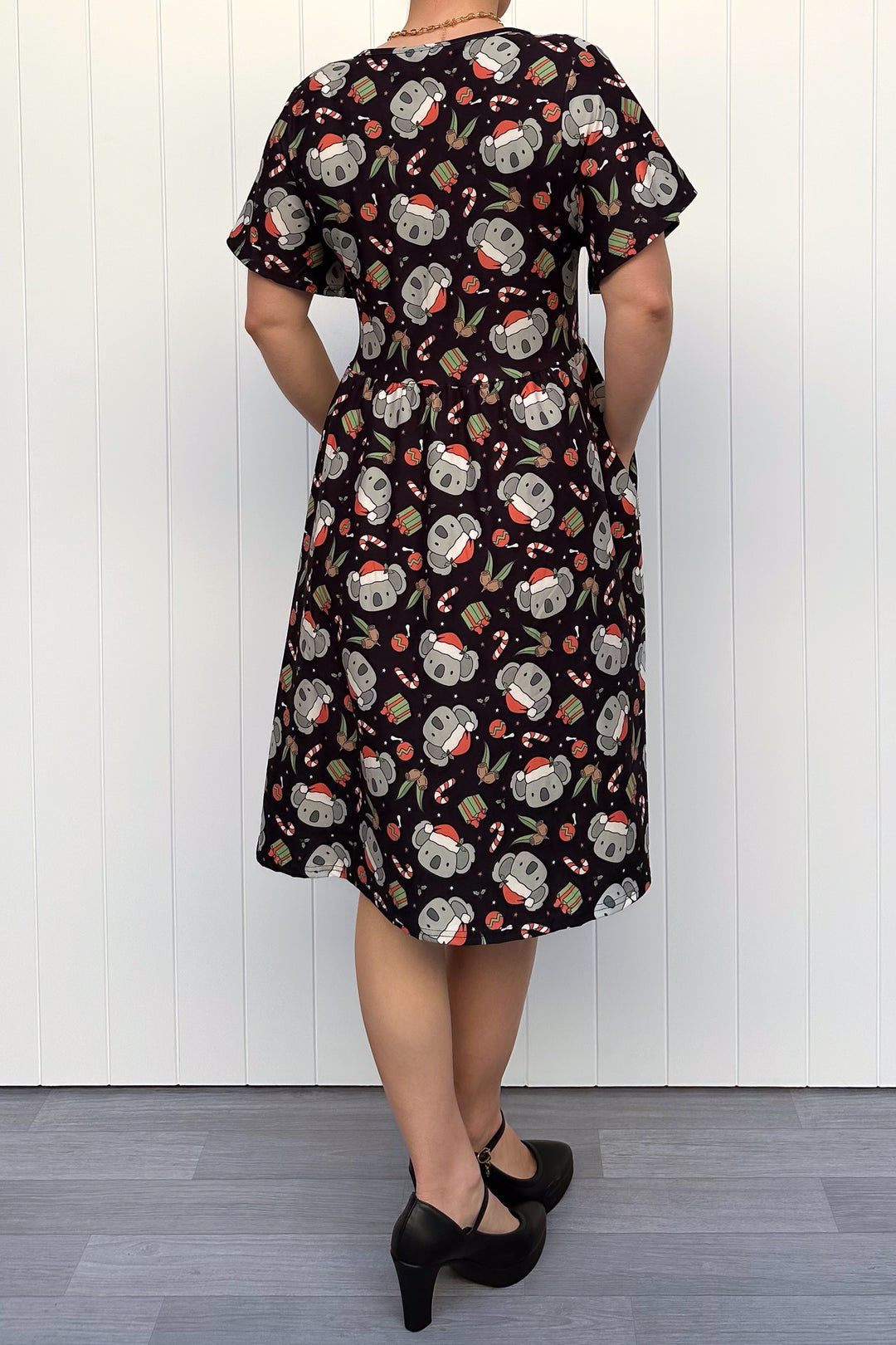 Person wearing a dress featuring a pattern of koalas wearing santa hats and candy canes on a black background, standing against a white wall.