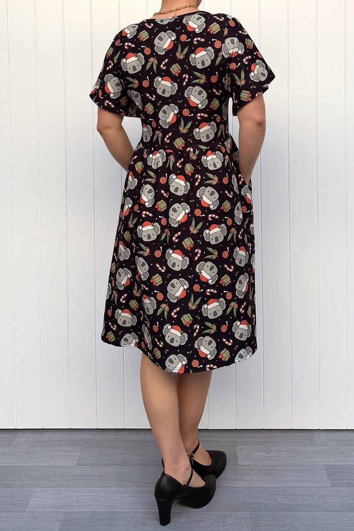 Person wearing a dress featuring a pattern of koalas wearing santa hats and candy canes on a black background, standing against a white wall.