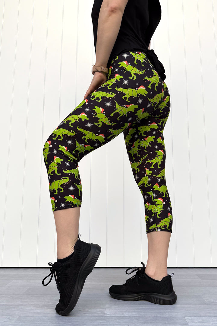 Person wearing capri leggings with green dinosaurs wearing santa hats on a black background.