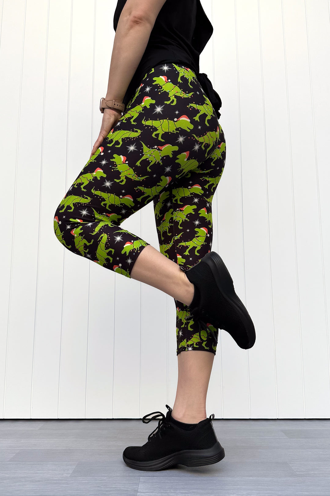 Person wearing capri leggings with green dinosaurs wearing santa hats on a black background.
