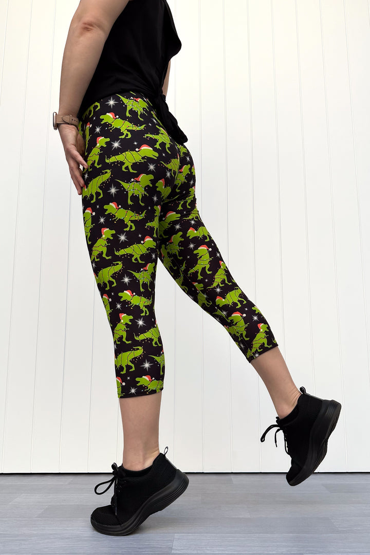 Person wearing capri leggings with green dinosaurs wearing santa hats on a black background.