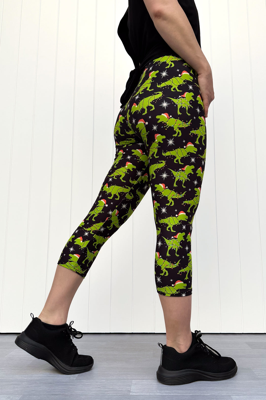 Person wearing capri leggings with green dinosaurs wearing santa hats on a black background.