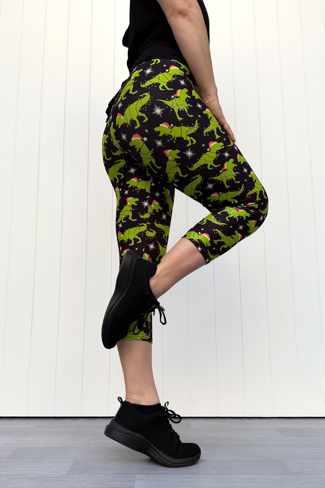 Person wearing capri leggings with green dinosaurs wearing santa hats on a black background.