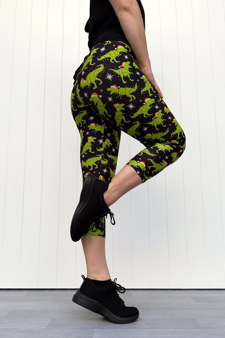 Person wearing capri leggings with green dinosaurs wearing santa hats on a black background.