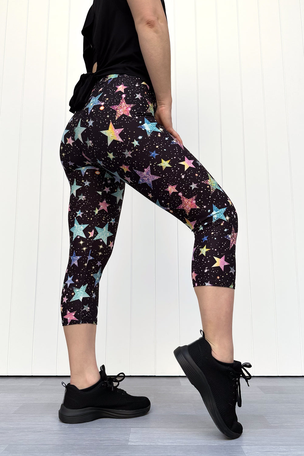 Person wearing black capri leggings with colourful sparkle-like star pattern, standing against a white wall.